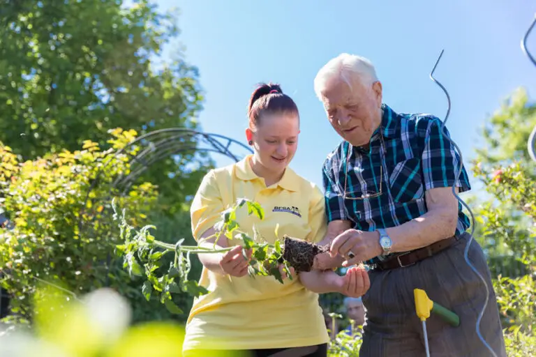 Senior und Mitarbeiterin pflanzen gemeinsam im Garten eine Tomate ein.
