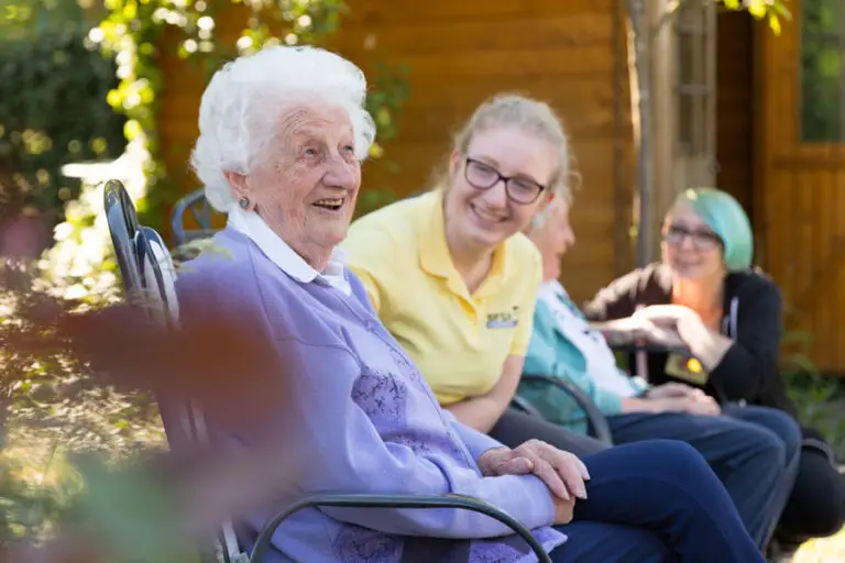 Seniorin sitzt lächelnd mit Betreuungskräften im Garten und genießt die Sonne.