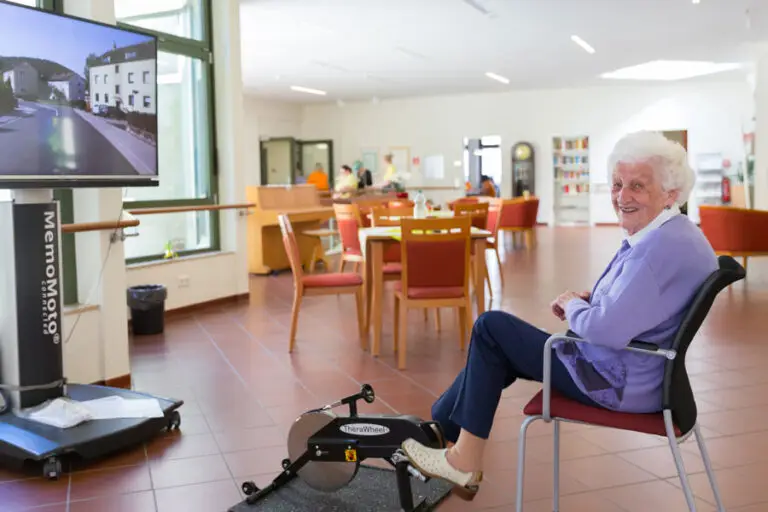 Seniorin trainiert auf einem Bewegungstrainer vor einem Bildschirm im Gemeinschaftsraum.