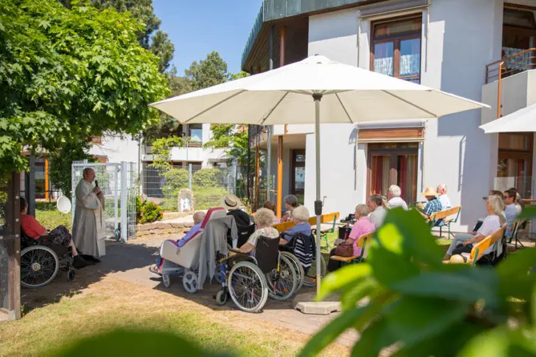 Gottesdienst im Garten, Bewohnerinnen und Bewohner sitzen im Schatten unter Sonnenschirmen.